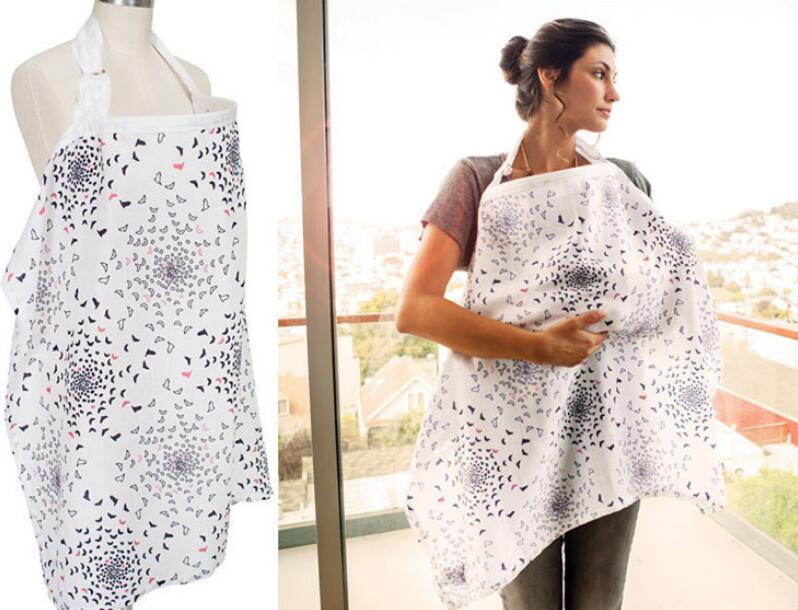 Woman using a patterned nursing cover while standing on a balcony. The white cover features a design with clusters of small, colored shapes. A mannequin displays the same cover.