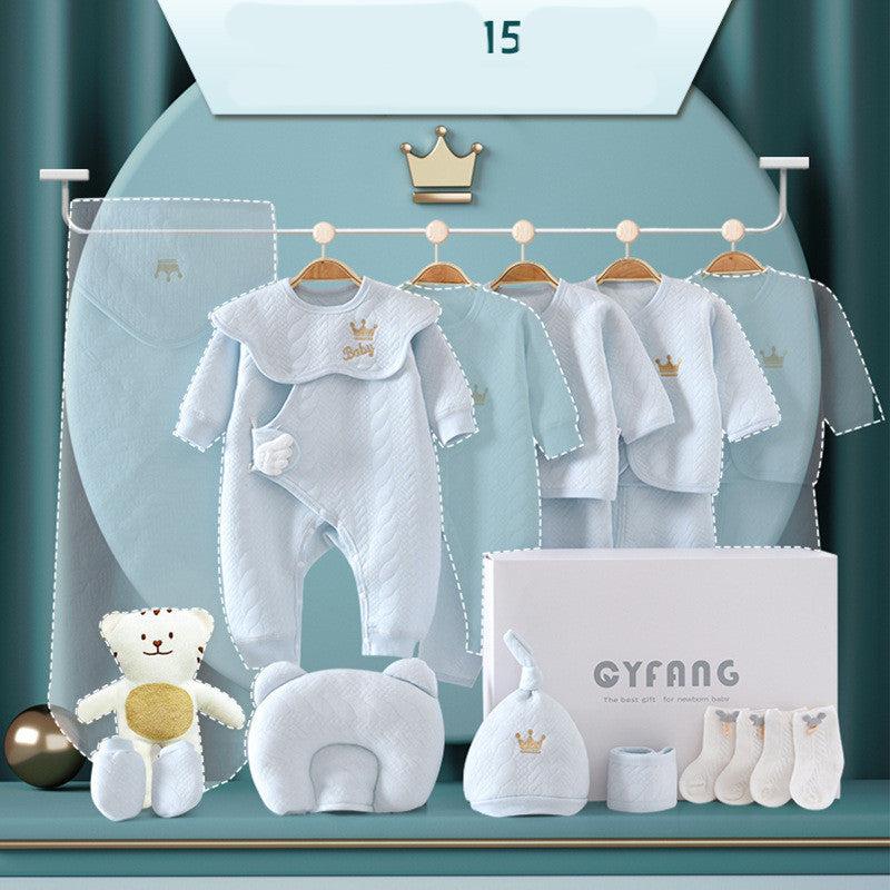 Baby gift set featuring light blue clothing with gold crown detail, a plush teddy bear, a small pillow, hat, box, and socks, displayed on a teal background.
