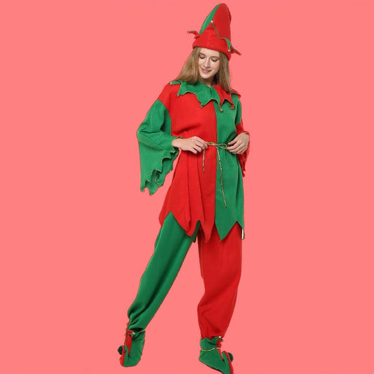 Person in a festive red and green elf costume with a pointed hat and decorative belt, standing on a solid pink background, smiling gently at the camera.
