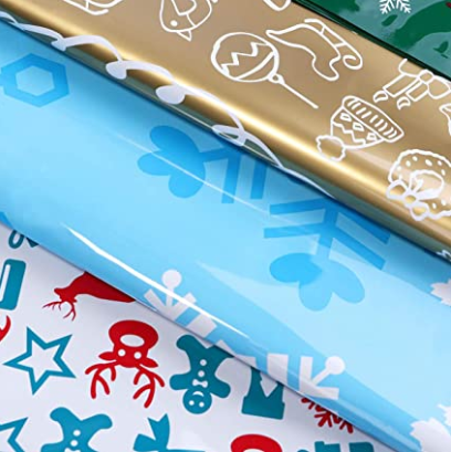Stacks of colorful wrapping paper, including gold with white holiday icons, light blue with large snowflakes, and white with red and blue festive symbols like stars and trees.