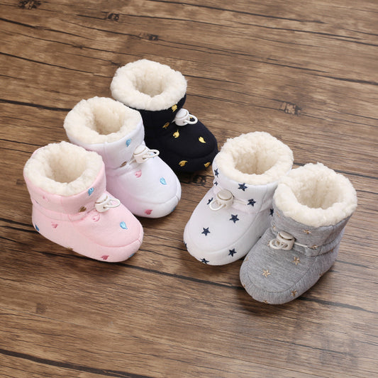 Five pairs of cozy baby booties with soft, fluffy inner lining displayed on a wooden floor. They come in pink, navy, white, and gray, each with star or heart patterns.
