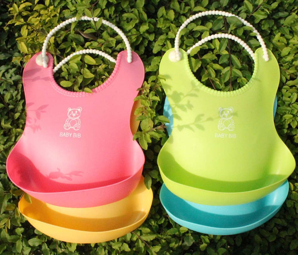 Two silicone baby bibs hang on a leafy background; one is pink with a yellow pocket, the other green with a blue pocket. Both have cartoon bears and "BABY BIB" printed on them.