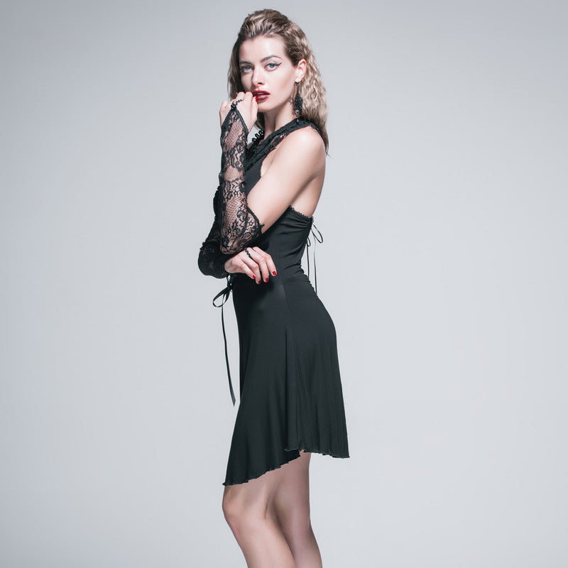 Woman in a black dress with lace gloves, standing in profile. She has bold makeup, long earrings, and red nails, striking a thoughtful pose against a gray background.