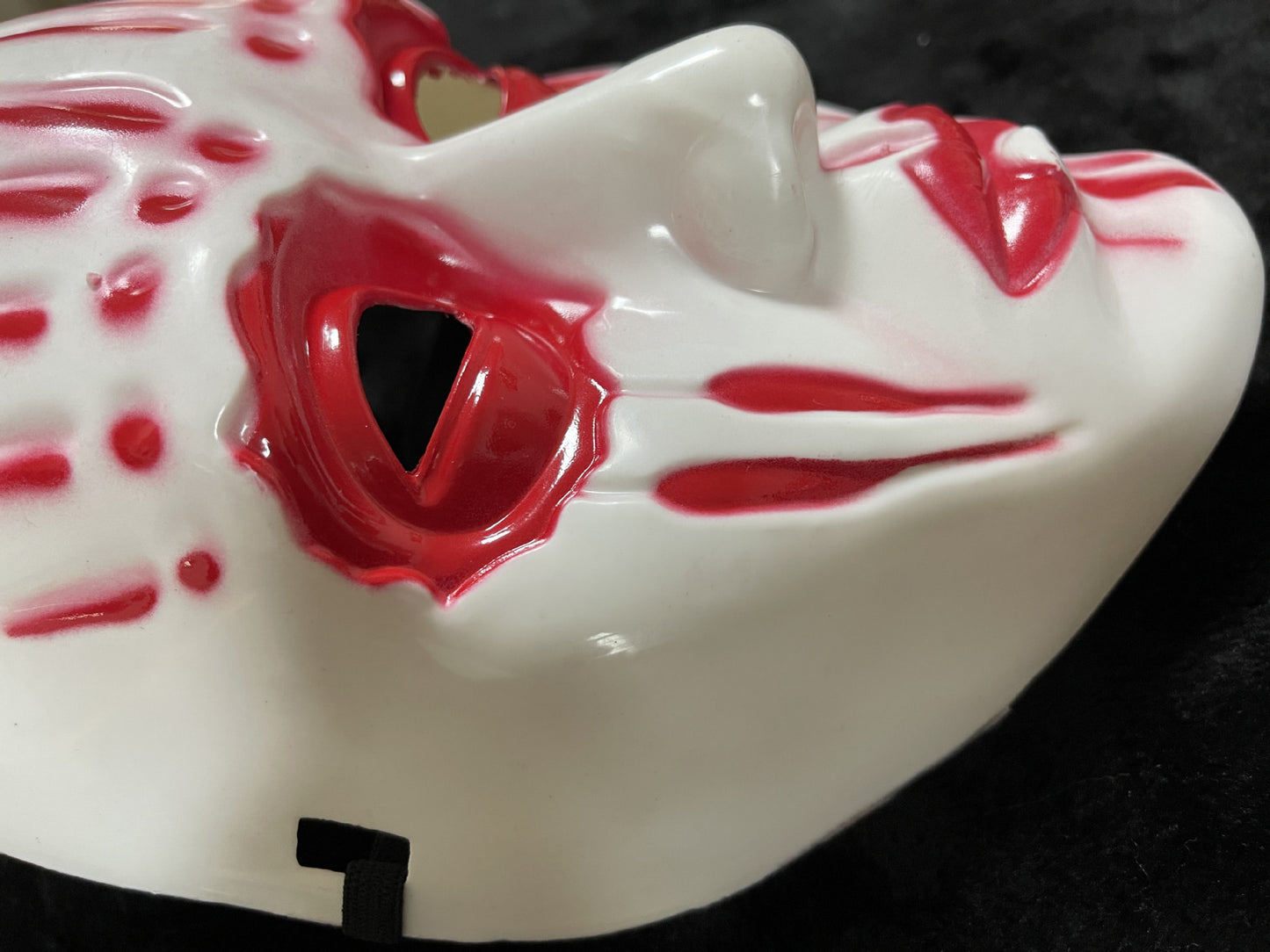 A white mask with red streaks and patterns, including eye outlines and lips, on a black background. The mask's dramatic design is emphasized by the contrast with the dark surface.