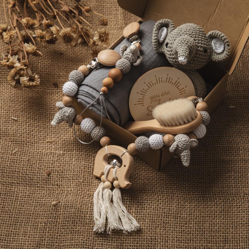 A cardboard box contains a handmade crocheted elephant, a wooden hairbrush, a soft cloth, and a string of wooden and crocheted beads on burlap fabric with dried flowers nearby.