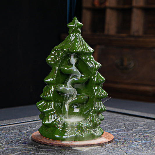 Green ceramic incense holder shaped like a Christmas tree, with smoke spiraling down its tiers on a textured surface. A cozy interior setting with a wooden backdrop.
