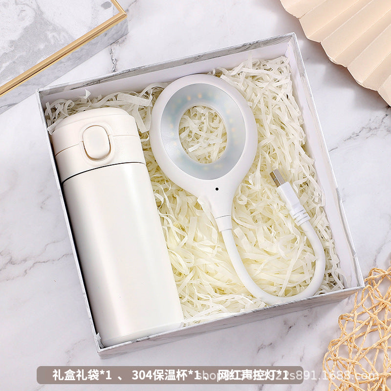 White thermos and circular LED light with USB cable in a box filled with white shredded paper, placed on a marble surface, partially covered by a beige fan.