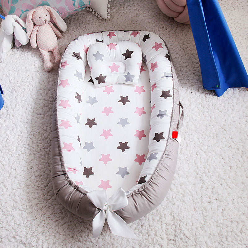 Soft baby nest with star pattern in pink, grey, and brown. It sits on a textured white carpet, next to plush toys and a blue fabric detail.