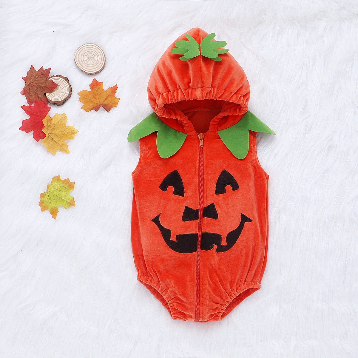 Orange pumpkin-themed baby romper with a happy jack-o'-lantern face, green leaf collar, and hood, placed on a white furry background alongside autumn leaves and wooden circles.