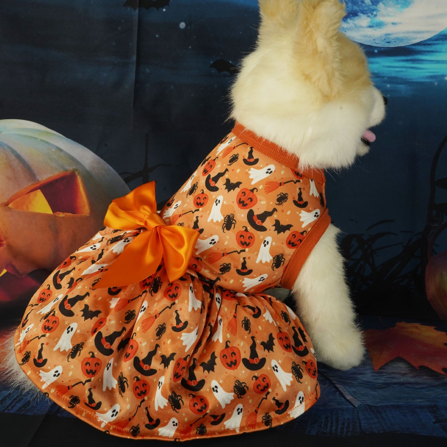 A fluffy dog wearing an orange Halloween-themed dress with ghosts and pumpkins, featuring a large bow, sits in front of a spooky backdrop with a carved pumpkin and moon.