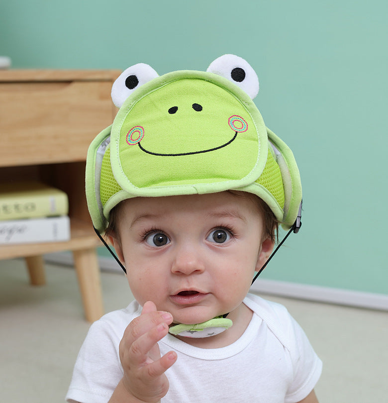 Baby wearing a green frog-themed helmet with wide eyes and a curious expression, seated in front of a pastel mint wall and wooden furniture with books on top.