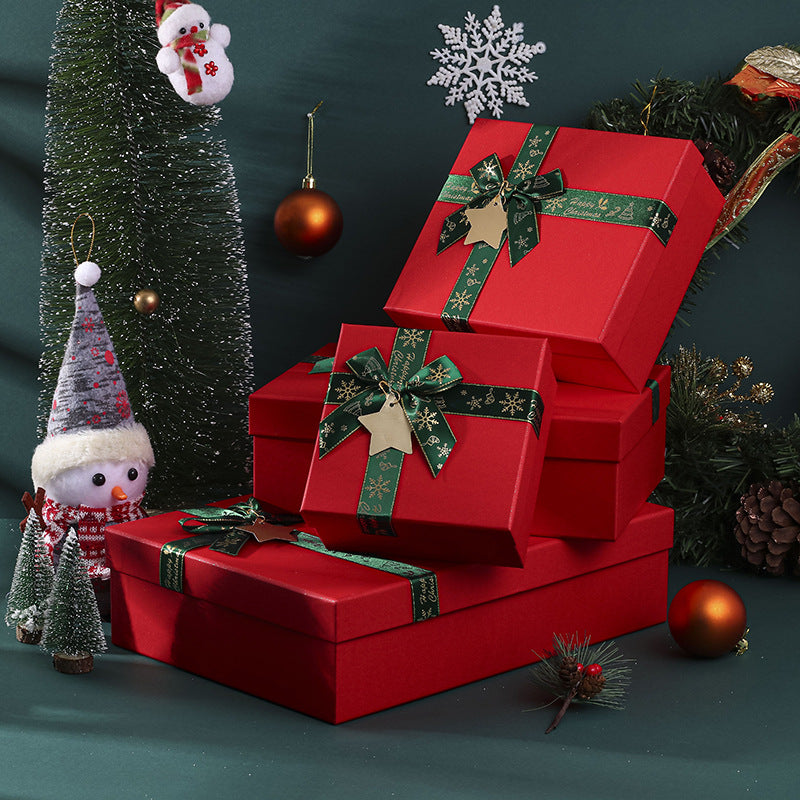 Stacked red gift boxes with green ribbons, a small snowman, Christmas trees, a hanging Santa, and ornaments decorate a festive holiday setting with pine cones and greenery.