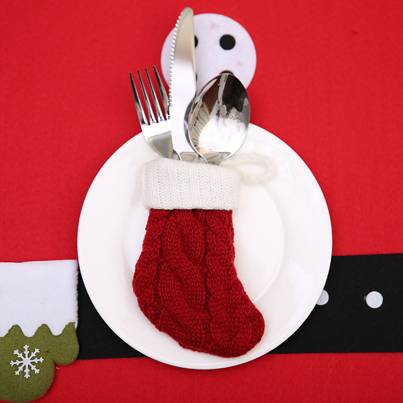 A red knitted Christmas stocking holds silver cutlery on a white plate, set against a red background with a black belt design, resembling Santa's outfit.