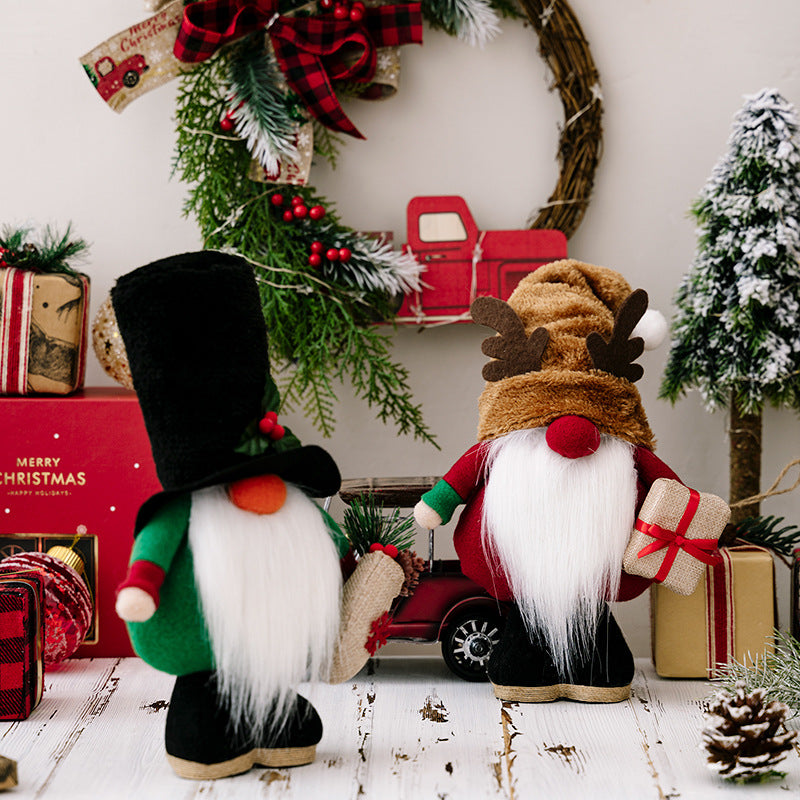 Two festive gnome figurines with white beards and tall hats stand among wrapped gifts. A Christmas wreath and small red truck decorate the background with snowy trees.