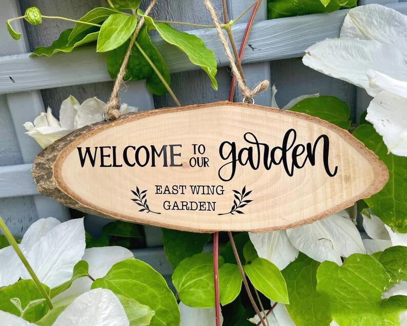 Wooden garden sign saying "Welcome to Our Garden - East Wing Garden" hangs on a lattice, surrounded by green leaves and white flowers.