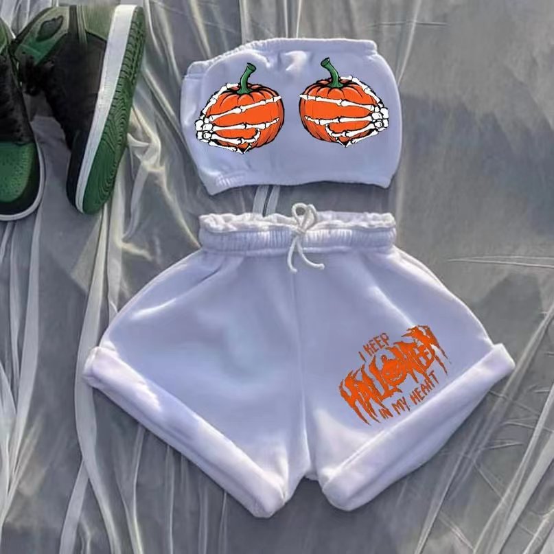White two-piece outfit featuring a tube top with orange pumpkins and skeleton hands design, paired with matching shorts displaying "I Keep Halloween in My Heart" in an orange, spooky font. Black and green sneakers lie beside the outfit on a gray surface.