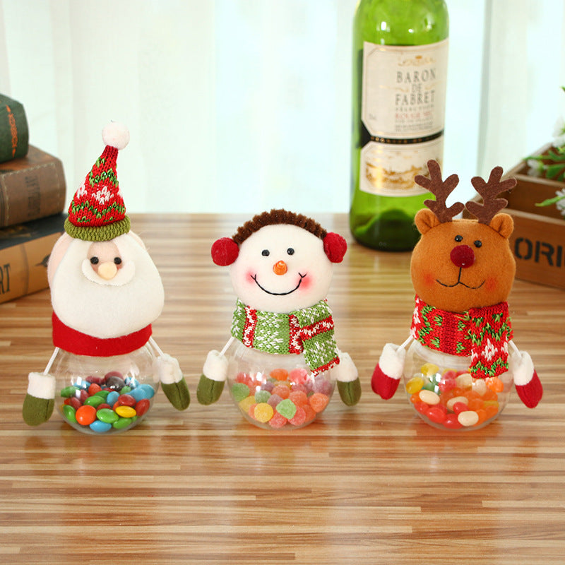 Three festive candy jars on a table, shaped like Santa, a snowman, and a reindeer, each filled with colorful candies. A wine bottle and books are in the background.