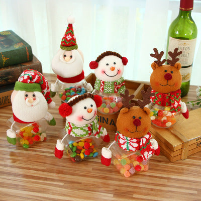 Festive plush figures of Santa, snowmen, and reindeer hold jars filled with colorful jelly beans. They wear holiday hats and scarves, set on a wooden table beside books and a wine bottle.