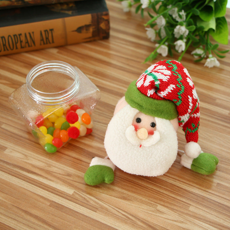 A plush Santa figure with a red and green hat sits on a wooden surface beside a glass jar of colorful candies. Behind it, a potted plant and books add a cozy touch.