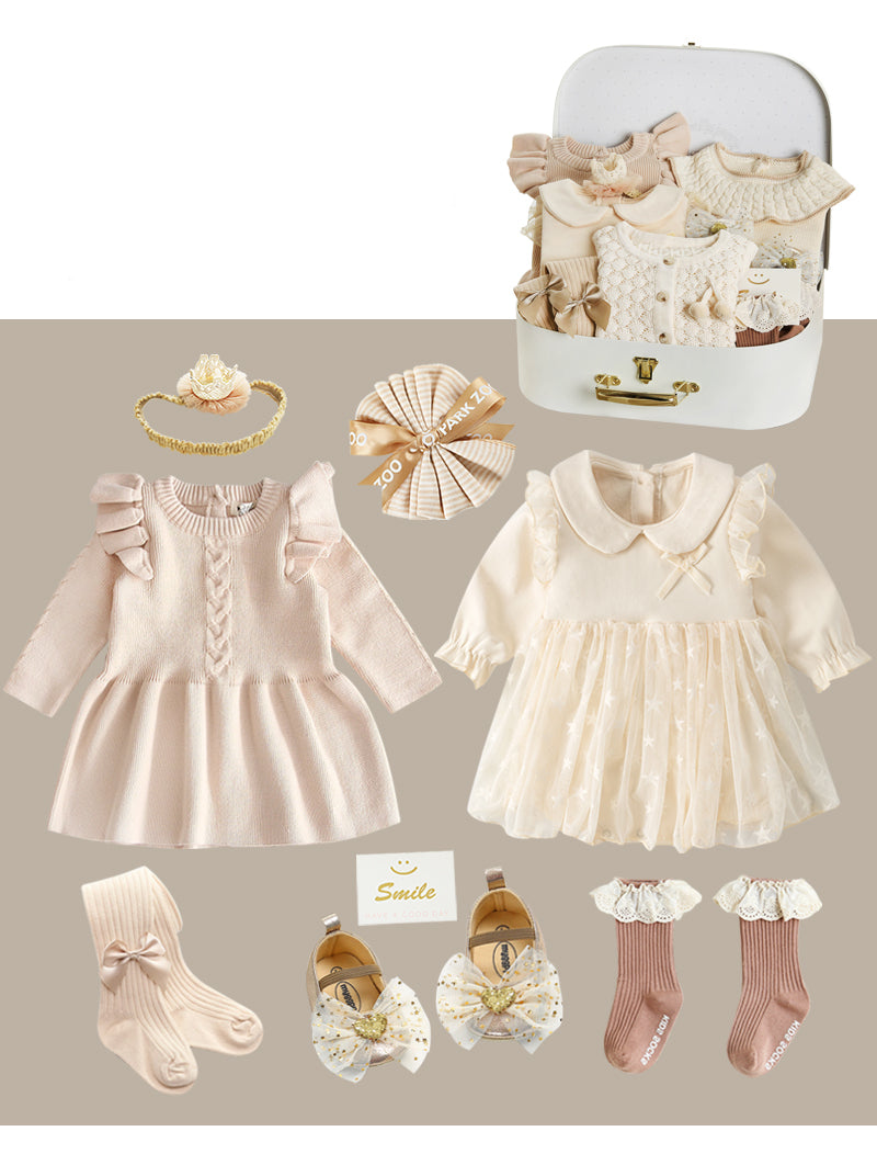 A collection of baby girl clothes and accessories in soft beige tones, featuring two dresses with ruffles, patterned socks, shoes with bows, hairbands, and a small trunk.