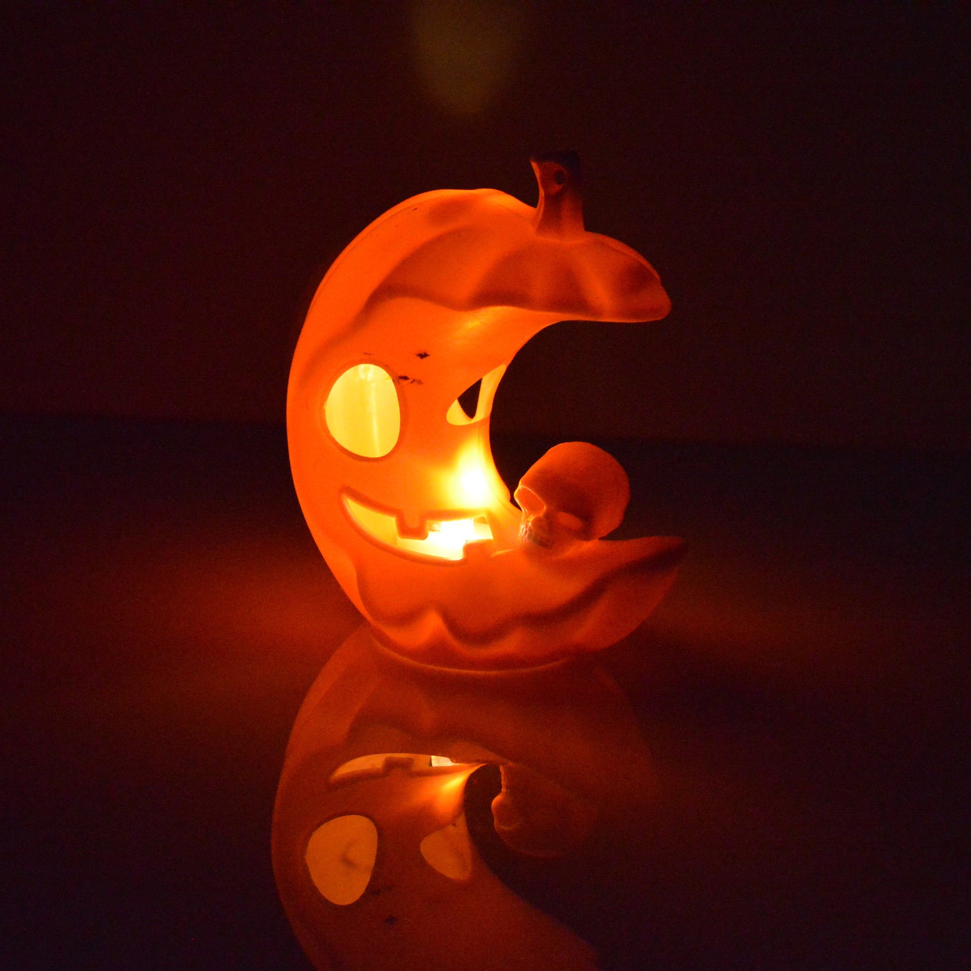 Glowing ceramic pumpkin decoration shaped like a crescent moon with a smiling face and small skull, illuminated by candlelight, casting a reflection on a dark surface.