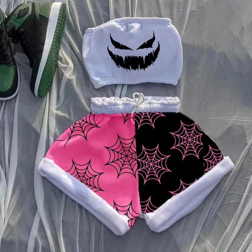 A stylish outfit consists of a white tube top with a menacing black face graphic and matching shorts. The shorts are divided into pink and black sections, each adorned with spiderweb patterns. Black and green sneakers are partially visible in the corner, all laid out on a sheer fabric background.