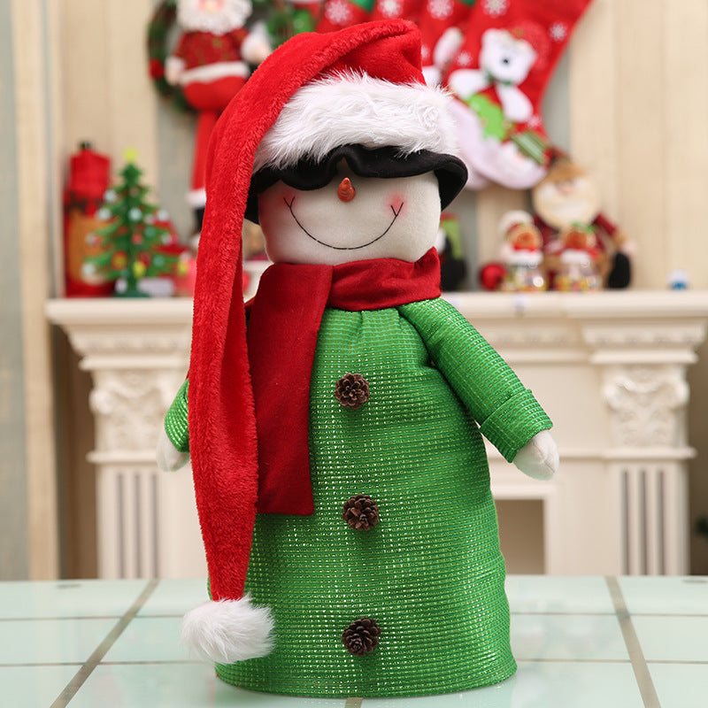 Smiling snowman doll with a red Santa hat and scarf, wearing a green textured coat adorned with pinecones. Festive background features Christmas decorations on a mantle.