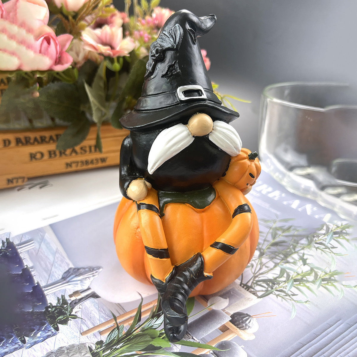 A whimsical gnome figurine wearing a tall black witch hat sits on a pumpkin. The gnome has a white mustache and holds a small jack-o'-lantern. Flowers and glassware are in the background.