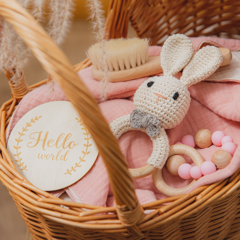 A wicker basket contains a pink cloth, wooden baby brush, crocheted bunny rattle, pink and wooden bead teether, and a round plaque saying "Hello world" in gold letters.