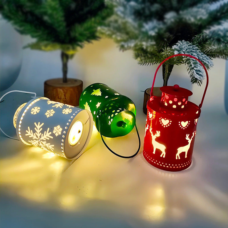 Festive lanterns on a table: a white one with snowflakes, a green one with stars, and a red one with reindeer designs, casting warm glows. Snowy trees in the background.