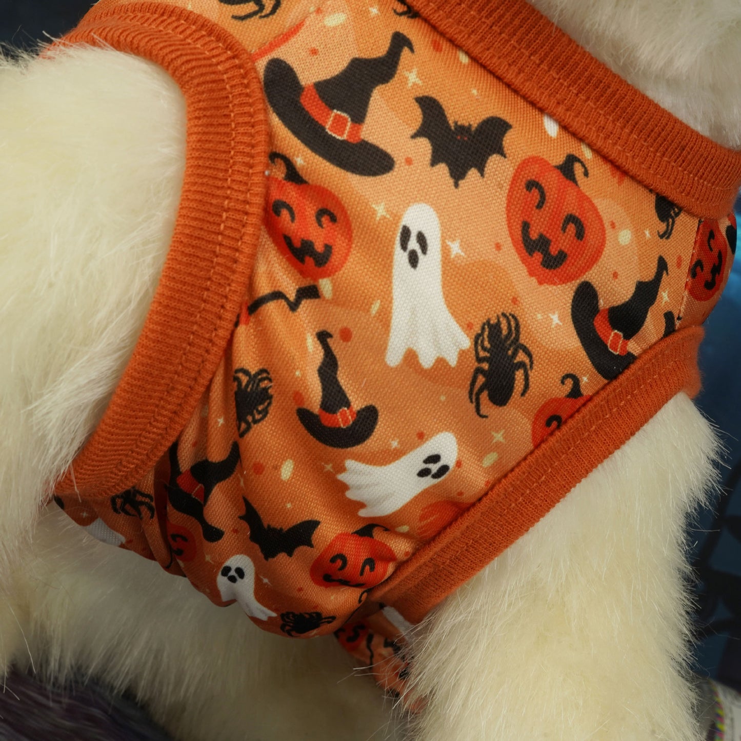 Close-up of a pet wearing an orange Halloween-themed outfit featuring ghosts, pumpkins, witch hats, bats, and spiders. The fabric is trimmed with orange edging.