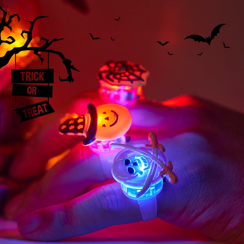 Close-up of a hand wearing Halloween-themed light-up rings, including a pumpkin, spiderweb, and skeleton. Background features "Trick or Treat" sign and silhouetted bats.