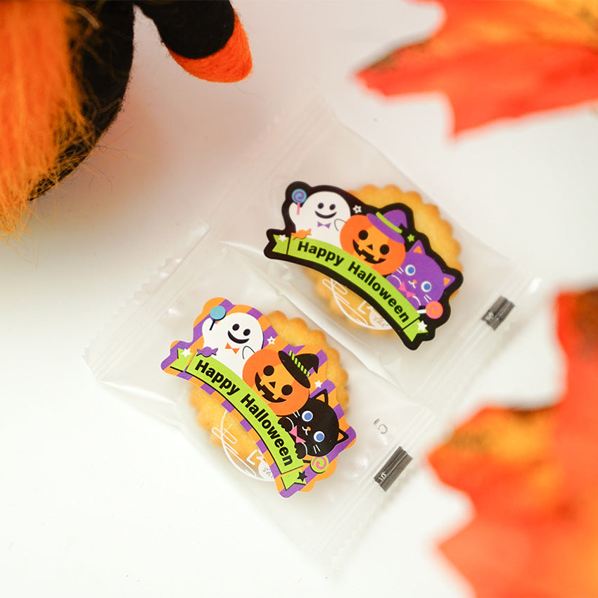Two packaged cookies with "Happy Halloween" stickers featuring a ghost, pumpkin, and black cat. The cookies are surrounded by Halloween-themed decorations, including an orange and black plush and autumn leaves, creating a festive seasonal atmosphere against a white background.