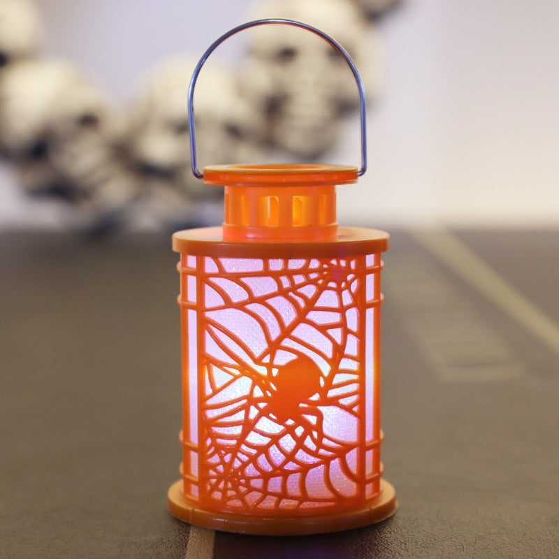 A glowing orange lantern with a spiderweb pattern stands on a flat surface. The lantern's light softly illuminates the intricate web design. A thin handle is attached to the top. In the blurred background, a garland of white skulls is visible, enhancing the Halloween-themed ambiance.
