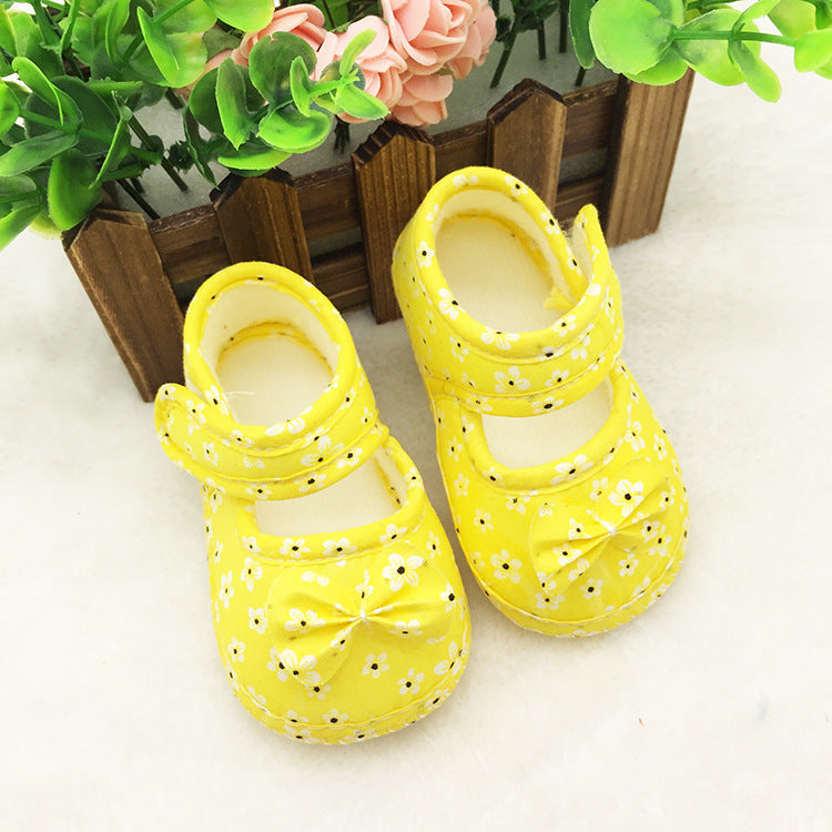 Yellow baby shoes with white daisies and a bow accent, placed on a soft surface next to a small wooden fence and green foliage with pink flowers.