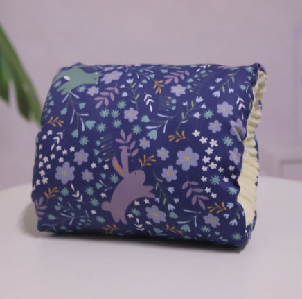 A plush pillow with a dark blue fabric, adorned with purple rabbits and light blue flowers, displayed on a white surface. The background is softly blurred with hints of green foliage.