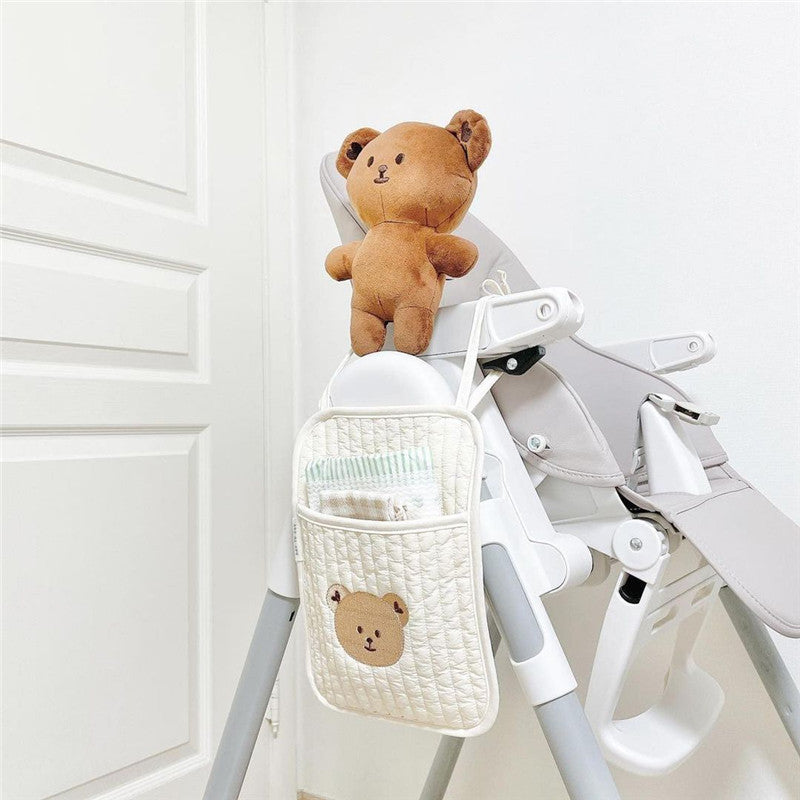 A high chair with a brown teddy bear on top and a quilted pocket on the side holding baby essentials. A bear face decorates the pocket. Background shows a white door and wall.