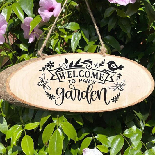 Oval wood sign with "Welcome to Pam's Garden" in black letters, decorated with floral designs, a bird, and a bee, hangs amid vibrant green leaves and purple flowers.