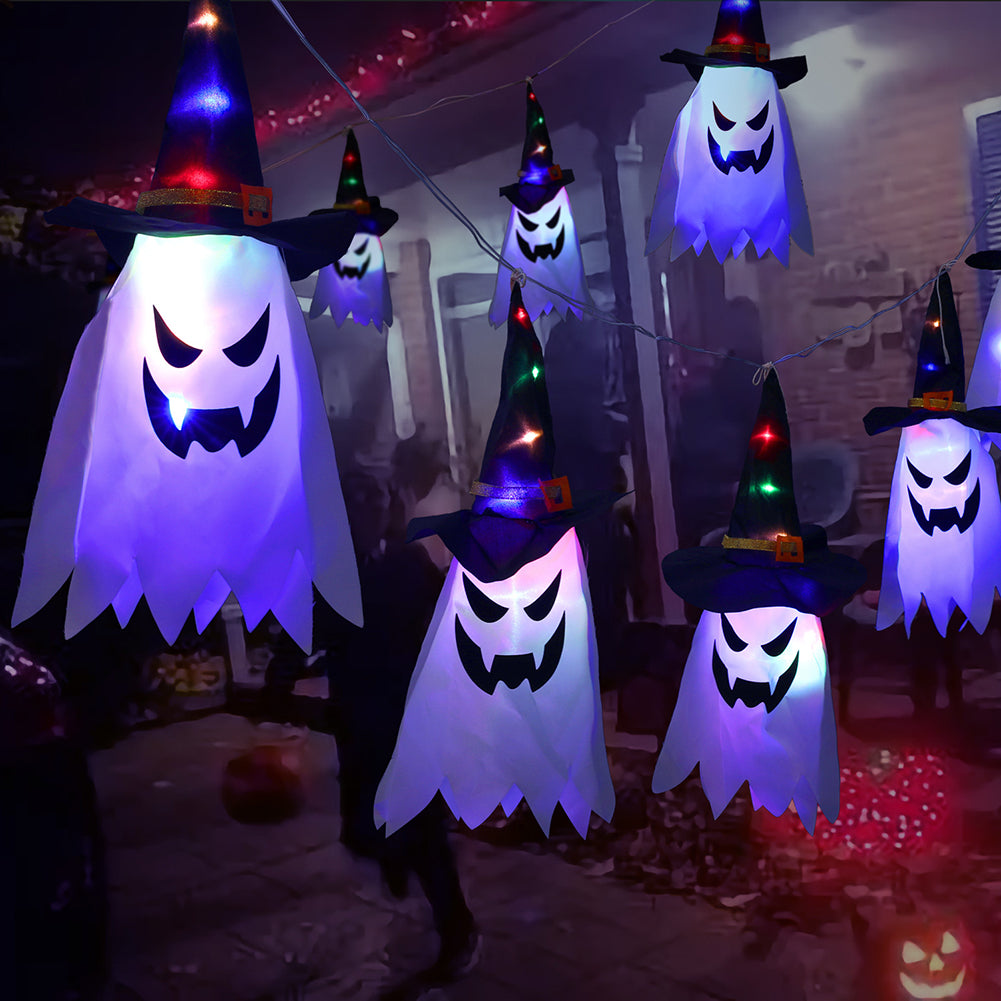 Glowing ghost decorations with witch hats hang in a festive, dimly-lit outdoor setting. The spooky faces are illuminated, adding to the Halloween ambiance.