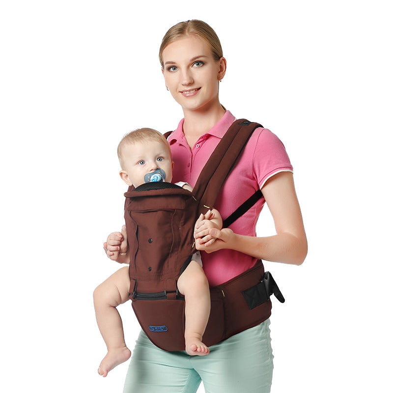 A woman in a pink shirt holds a baby in a brown front-facing carrier. The baby, with a pacifier, looks at the camera. The background is white and plain.