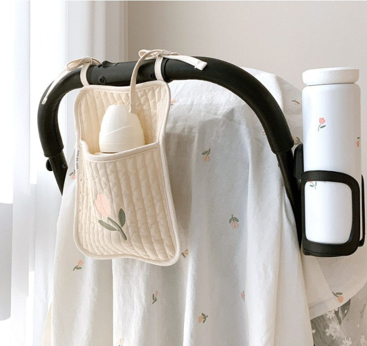 A stroller handle with a quilted beige pouch holding a bottle, and a white thermos attached. The stroller cover is white with a subtle floral pattern in a softly lit room.