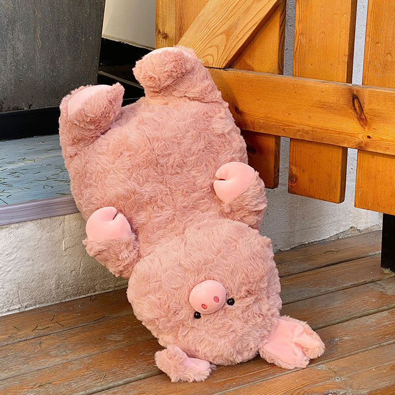 A large, fluffy pink stuffed pig toy is upside down, resting against a wooden bench on a wooden floor. Its eyes, snout, and ears are visible, adding to its playful appearance.