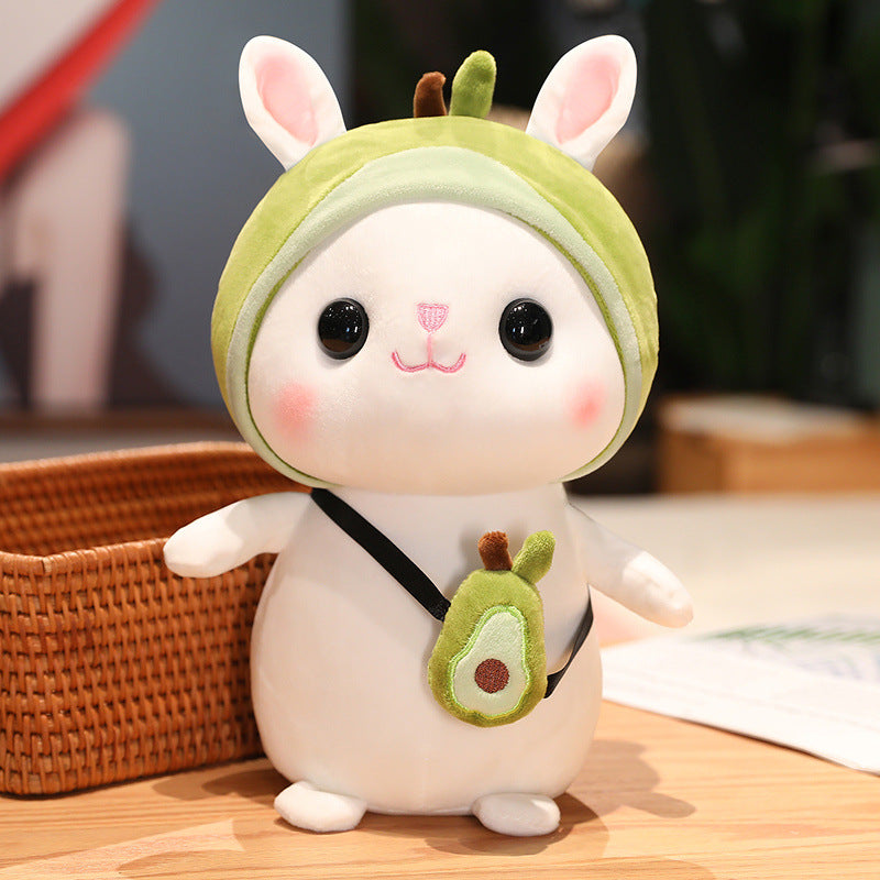 Cute plush toy of a white animal with rosy cheeks, big eyes, wearing a green apple hat and sling bag. It stands on a light wooden surface next to a wicker basket.