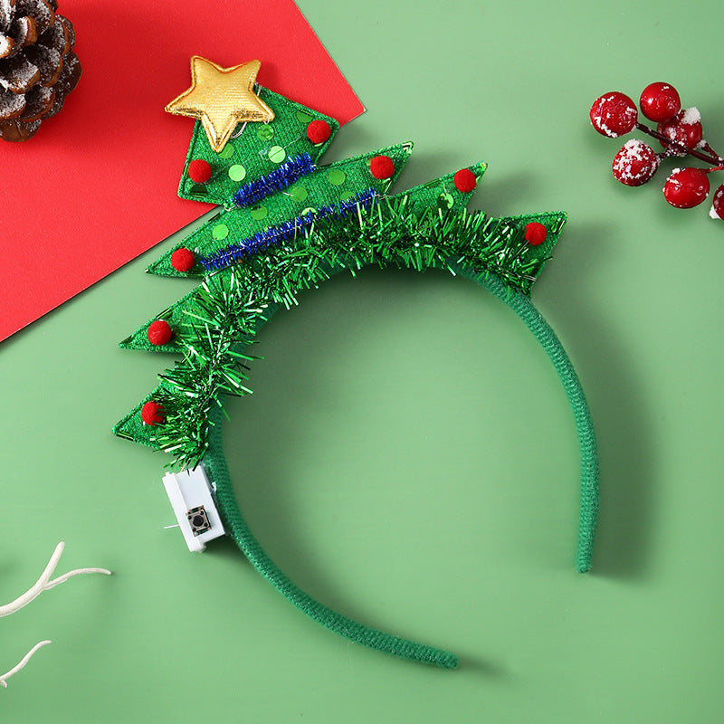 Green headband decorated as a Christmas tree with a gold star, red dots, and tinsel on a green backdrop. Nearby, red berries, pine cone, and antler decor add to the festive theme.