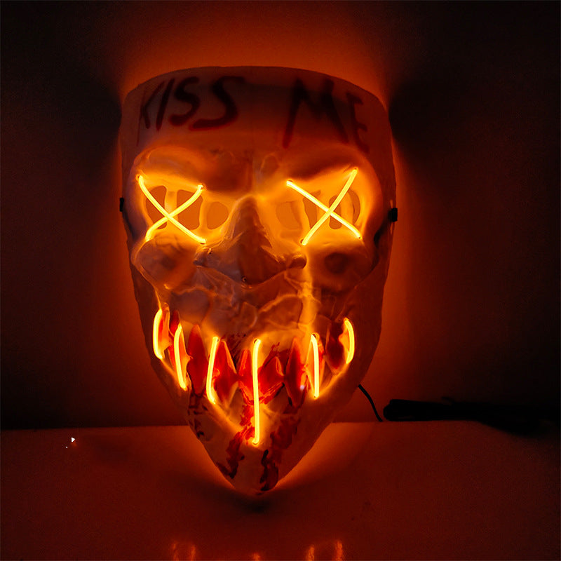 Glowing mask with "Kiss Me" inscribed. Features neon X eyes and jagged mouth, emitting an eerie orange light against a dark background.
