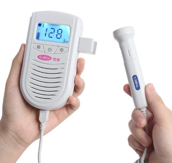Two hands holding a fetal Doppler device; one hand holds the monitor with a digital screen displaying numbers, the other holds the probe labeled "3 MHz" with a connecting wire.
