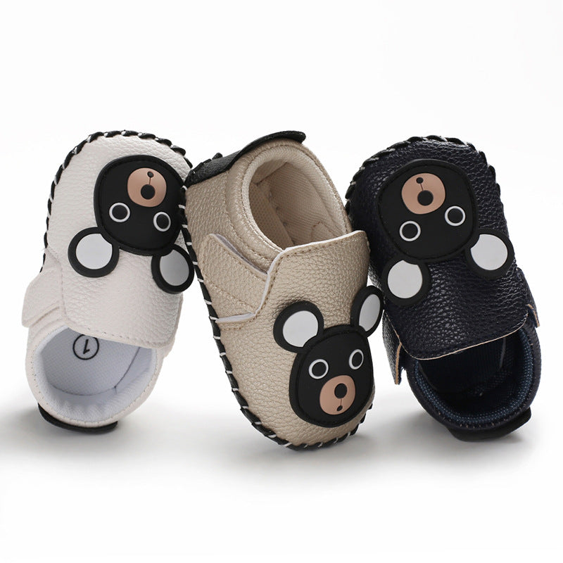 Three baby shoes with cute bear face designs in white, beige, and navy colors. Each shoe features black bear details and a soft, textured surface, arranged upright and side by side.