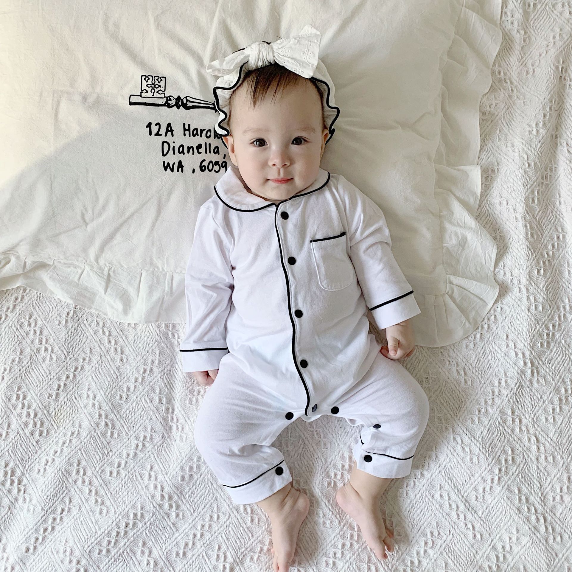 A baby in white pajamas with black trim and a matching headband lies on a patterned blanket, looking up with a slight smile. The pillow has whimsical text and a drawing.