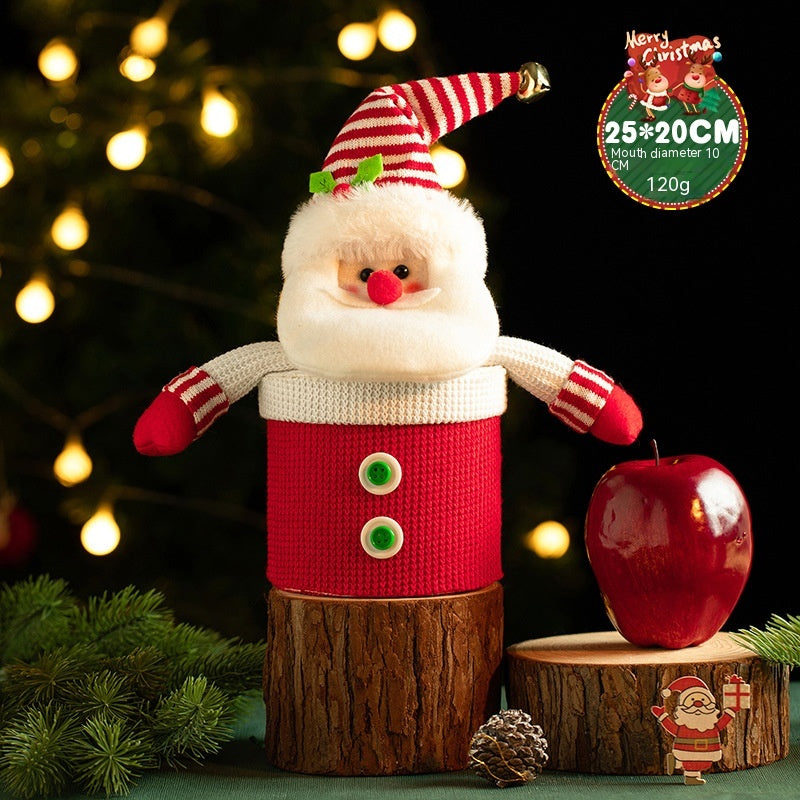 Festive Santa Claus figure with a knitted red outfit and striped hat, set on a wooden stand beside a shiny red apple, pine sprig, and cone, with blurred lights in the background.