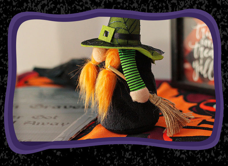 A small plush witch figurine with an orange beard, wearing a green and black hat and striped green sleeves, holds a tiny broom. It is displayed on a colorful tablecloth with blurred background elements, including framed text. The image is bordered by a wavy purple frame.